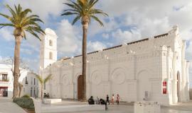 Se ve el edificio blanco con sus murallas y su gran torre y en la plaza 2 palmeras que se elevan hasta la misma altura de la torre en un día soleado con nubes blancas encima del edificio