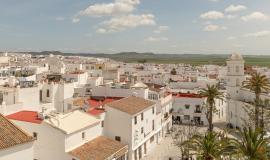 Se observa una vista aérea del pueblo blanco de Conil con la plaza Sta Catalina y sus palmeras y al fondo un valle verde de campo 