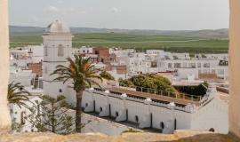 Se aprecia a través de una ventana de piedra desde una torre un edificio blanco amurallado con una palmera verde en la plaza y al fondo el pueblo lleno de casitas blancas y un prado verde más al horizonte en un día soleado