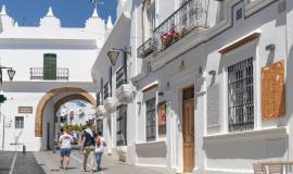 Se ven 3 personas de espaldas caminando a través de una calle rodeada de casas blancas y al final de la misma un arco para  pasar a una plaza