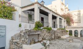 Se ve un patio con una fuente antigua de piedra y encima una casa blanca con flores rojas colgando de sus paredes