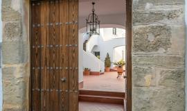 Se ve una fachada en primer plano de piedra con un porto de madera que esta abierto y se observa en el interior un patio lleno de macetas con flores