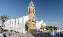 Se ve una iglesia a media distancia en un día soleado en el que predominan sus muros blancos y una torre de piedra que parece alzarse hasta el cielo