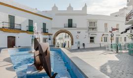 Se ve un patio rodeado de casas blancas y en el medio una fuente con 3 peces de bronce y un agua azulada en un día soleado