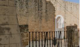 Se observa un patio a través de unas rejas y al fondo una torre que se alza sobre la muralla en un día soleado