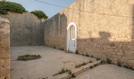 Se aprecia un patio amurallado, con árboles asomando al fondo y una puerta blanca en el medio del patio, con medias sombras y sol