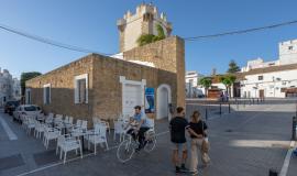 Se aprecia un hombre en bicicleta y una pareja paseando por la sombra de una calle en la que se pueden observar sillas y mesas blancas de una bar al lado de una edificación con muralla antigua