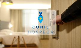 Se puede obsevar una puerta entre abierta enseñando una habitación de hotel a través de ella con el símbolo de un icono en medio que indica Conil Hospeda con el dibujo de un pescado hacia abajo